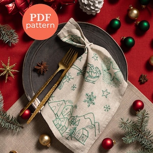 Puede incluir: Una mesa festiva navideña con una servilleta de lino con un diseño de pueblo invernal bordado en verde, cubiertos dorados y adornos rojos y verdes. La servilleta está sobre un plato gris oscuro. El texto "PDF pattern" está en un círculo.