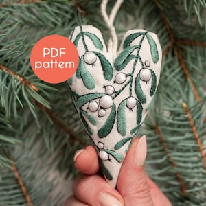 May include: A white heart-shaped ornament with green embroidery of mistletoe leaves and white berries. The ornament is hanging from a string and is held in a hand. The text "PDF pattern" is on a red circle in the background.