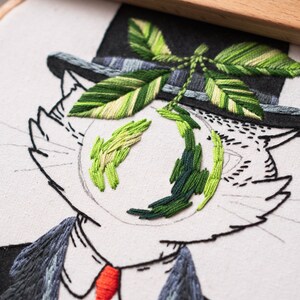 May include: Embroidered artwork of a cat in a top hat and suit, with a red tie. The cat's eyes are stitched with green thread, and a green leaf design is above the cat's head. The embroidery is on a white fabric.