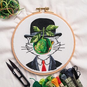 May include: An embroidered hoop art piece depicting a cat in a bowler hat, suit, and red tie, with a green apple as a face. The apple and leaves are intricately stitched. Embroidery floss and scissors are visible.