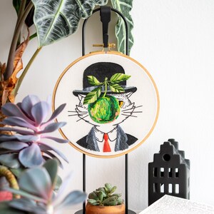 May include: Embroidered artwork in a wooden hoop depicts a cat in a bowler hat, suit, and red tie, with a green apple replacing its face. The cat is set against a white background, mounted on a black metal stand.