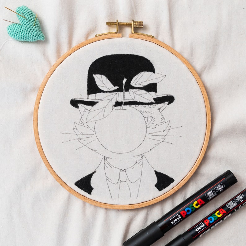 May include: An embroidery hoop featuring a cat design in a bowler hat and apple, inspired by Magritte. The design is in black on white fabric. Two black Uni Posca markers are next to the hoop. The hoop is made of wood.