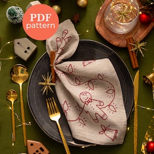 Pode incluir: Uma mesa festiva de Natal com um guardanapo cinza com um desenho de homem de gengibre, talheres dourados e enfeites de Natal. O guardanapo é dobrado e preso com um pau de canela. O texto "PDF pattern" é visível.