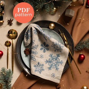 Könnte beinhalten: Festliche Tischdekoration mit einer beigen Leinen-Serviette, bestickt mit blauen Schneeflocken. Die Serviette liegt auf einem Teller mit goldenem Besteck. Die Szene ist mit Weihnachtsschmuck, Tannenzweigen und dem Text "PDF pattern" dekoriert.
