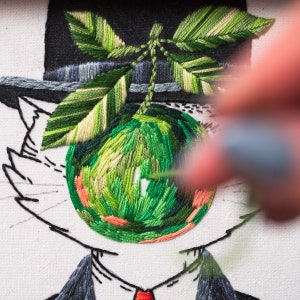 May include: Detailed embroidery of a cat in a hat and suit, with a green apple eye and leaves. The needlework uses green, orange, and black threads on a white canvas. The image shows a hand holding a needle, creating the embroidery.