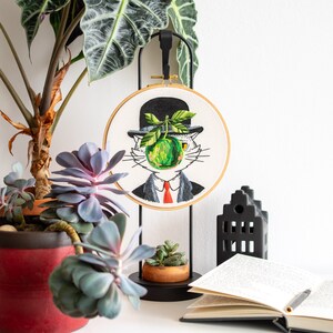 May include: An embroidered hoop art piece depicts a cat in a bowler hat with a green apple obscuring its face. The cat is dressed in a suit and red tie. The artwork is displayed on a black stand, with plants and a book nearby.