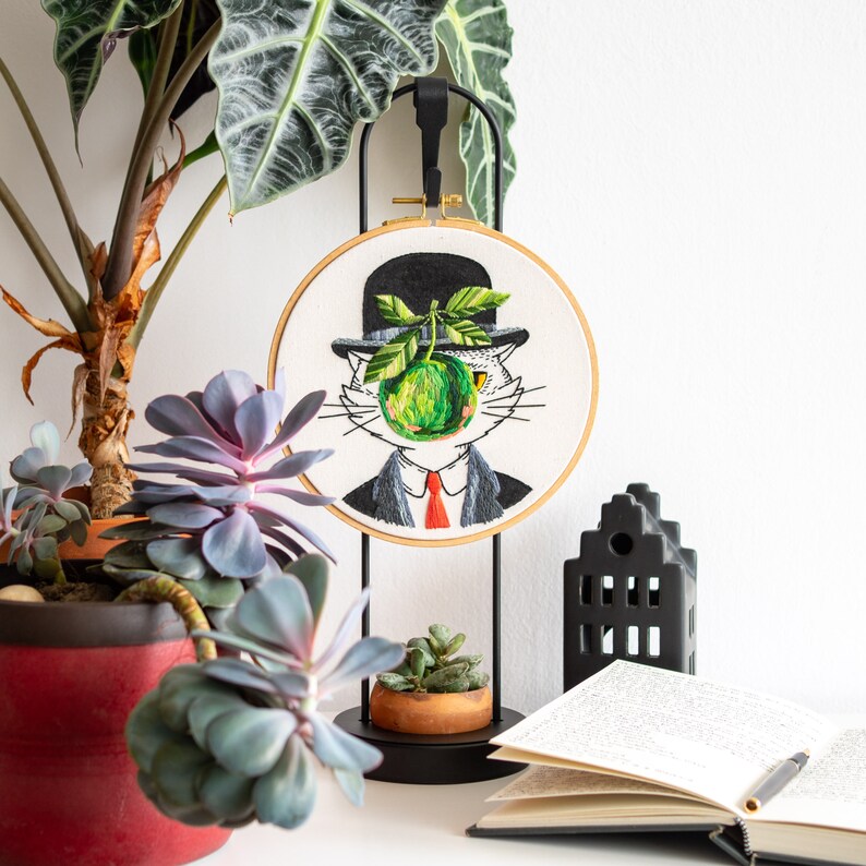 May include: An embroidered hoop art piece depicts a cat in a bowler hat with a green apple obscuring its face. The cat is dressed in a suit and red tie. The artwork is displayed on a black stand, with plants and a book nearby.