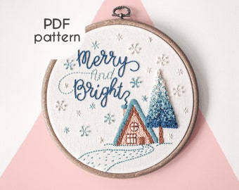 May include: Embroidered hoop art with a winter scene. The design features a small cabin, a blue and white ombre Christmas tree, and the text "Merry and Bright" in cursive script. The background is white with scattered snowflakes.