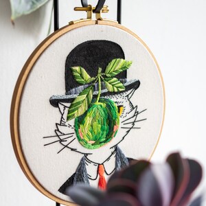 May include: Embroidered artwork in a wooden hoop depicts a cat in a bowler hat, with a green apple and leaves. The cat wears a suit and tie. The embroidery is on a white background, with a black ribbon and gold hardware for hanging.