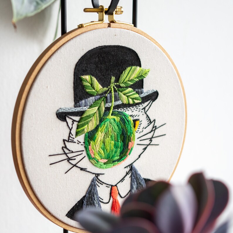 May include: Embroidered artwork in a wooden hoop depicts a cat in a bowler hat, with a green apple and leaves. The cat wears a suit and tie. The embroidery is on a white background, with a black ribbon and gold hardware for hanging.
