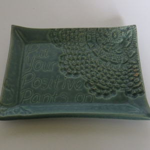 Puede incluir: Un plato cuadrado de cerámica color verde azulado con esquinas redondeadas. El plato presenta la frase "Put Your Positive Pants on" en relieve. Un patrón decorativo texturizado está en el lado derecho.