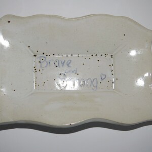 Pode incluir: Um prato de cerâmica artesanal, branco sujo, com um centro retangular e bordas onduladas. O prato é salpicado com pontos castanhos e apresenta as palavras "Brave and Strong" em escrita azul, com um pequeno símbolo de coração.