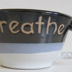 May include: A ceramic mug with a black top, light blue stripe, and white base. The word "breathe" is written in tan letters. The handle is a twisted rope design, transitioning from black to white.