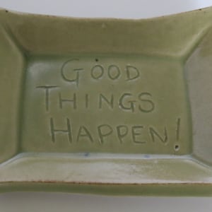 May include: A square, sage green ceramic dish with gently curved sides. The words "GOOD THINGS HAPPEN!" are etched into the center. The dish has a slightly textured surface and a light brown rim.