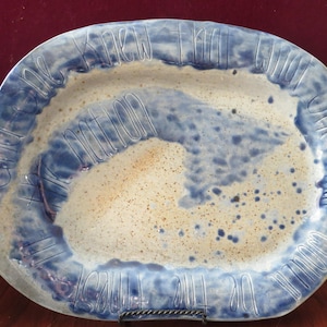 May include: Oval ceramic platter with a speckled off-white center and a blue rim. The rim features etched text and a wavy design. The platter is supported by a metal stand. The overall aesthetic is rustic and handmade.