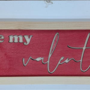 Puede incluir: Un letrero rectangular de madera con un marco de madera natural. El letrero tiene un fondo rojo con las palabras "... be my valentine" recortadas de la madera. Una pieza decorativa para el D&iacute;a de San Valent&iacute;n.