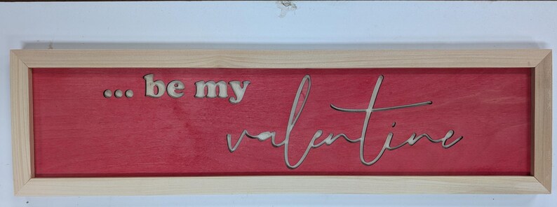 Puede incluir: Un letrero rectangular de madera con un marco de madera natural. El letrero tiene un fondo rojo con las palabras "... be my valentine" recortadas de la madera. Una pieza decorativa para el D&iacute;a de San Valent&iacute;n.