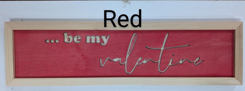 Handmade cypress frame with Baltic birch red background and natural Baltic birch letters reading ...be my Valentine. Be my is in printed letters and the valentine in script. The words are recessed into the wood. The wood grains is seen on all wood.