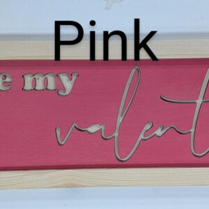 Handmade cypress frame with Baltic birch pink background and natural Baltic birch letters reading ...be my Valentine. Be my is in printed letters and the valentine in script. The words are recessed into the wood. The wood grains is seen on all wood.