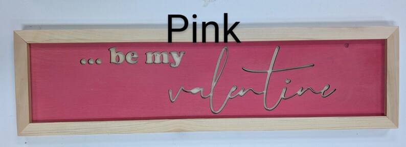 Handmade cypress frame with Baltic birch pink background and natural Baltic birch letters reading ...be my Valentine. Be my is in printed letters and the valentine in script. The words are recessed into the wood. The wood grains is seen on all wood.