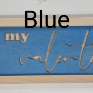 Handmade cypress frame with Baltic birch blue background and natural Baltic birch letters reading ...be my Valentine. Be my is in printed letters and the valentine in script. The words are recessed into the wood. The wood grains is seen on all wood.