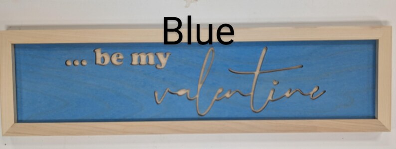 Handmade cypress frame with Baltic birch blue background and natural Baltic birch letters reading ...be my Valentine. Be my is in printed letters and the valentine in script. The words are recessed into the wood. The wood grains is seen on all wood.