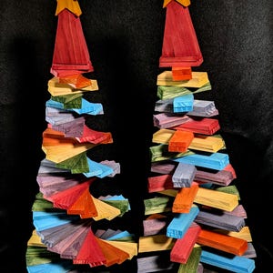 May include: Two decorative wooden trees featuring a spiral design made of stacked, multi-colored rectangular slats in shades of red, orange, yellow, green, blue, and purple. Each tree is topped with a solid yellow wooden star ornament.