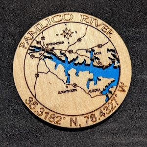May include: A round wooden coaster featuring a map of the Pamlico River with blue water and surrounding towns. The coaster includes the coordinates 35.3182° N, 76.4327° W and the text "Pamlico River".
