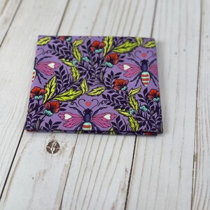 May include: A folded square of fabric with a repeating pattern of stylized insects, flowers, and foliage in shades of purple, red, yellow, and teal. The design features heart-shaped accents and a whimsical aesthetic.