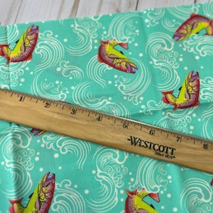 May include: Teal fabric featuring a repeating pattern of colourful fish and white wave designs. A wooden ruler with inch measurements is placed diagonally across the fabric. The ruler has the text "WESTCOTT Since 1872".
