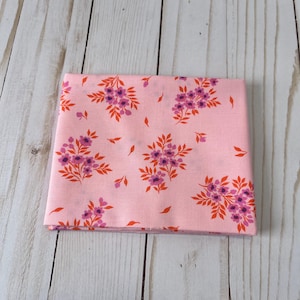 May include: Folded pink fabric with a floral pattern. The design features clusters of purple flowers and orange leaves scattered across the surface. The fabric is folded neatly, showcasing the pattern's repeat.