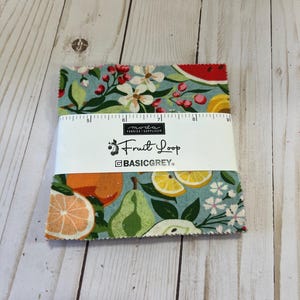May include: A fabric square with a blue background and a colorful fruit print. The fabric features lemons, oranges, pears, watermelons, and flowers. The fabric is labeled "Fruit Loop" and "BASICGREY."