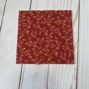 May include: A square piece of red fabric with a floral pattern. The fabric features small, light-colored flowers and leaves on thin stems. The edges of the fabric are cut with a zig-zag pattern. The fabric is placed on a light wood surface.