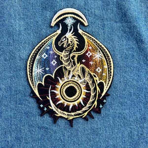 May include: Embroidered patch with a dragon design and a sun-like centre. The patch has a black background with gold, blue, and red accents. The dragon is gold, and the wings have a rainbow gradient. The patch is on a blue denim background.