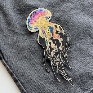 Puede incluir: Parche bordado de medusa en tonos arcoíris sobre fondo negro, cosido sobre unos shorts vaqueros grises. El parche presenta costuras detalladas y un diseño único, perfecto para añadir estilo a la ropa.