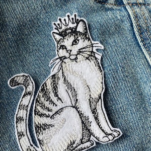 Puede incluir: Un parche de hierro bordado en blanco y negro de un gato con una corona. El gato está sentado con la cola enroscada alrededor de su cuerpo.