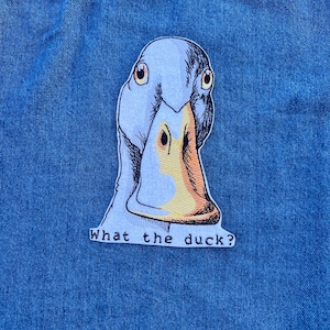 Puede incluir: Un parche bordado azul claro con la cara de un pato con ojos amarillos y un pico naranja. El texto "What the duck?" está debajo. El parche está sobre un fondo de mezclilla azul.