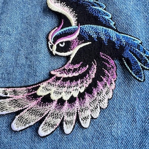 May include: Embroidered owl patch with detailed stitching in shades of blue, purple, and white on a black background. The patch is set against a blue denim fabric, showcasing the vibrant colors and intricate design of the bird.