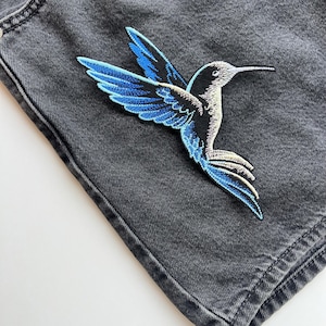 May include: A detailed embroidered hummingbird patch in blue and black, sewn onto grey denim. The patch depicts the bird in flight, with a long beak and detailed wing patterns. Suitable for adding a decorative element to clothing or accessories.