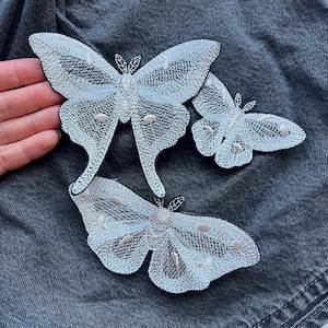 May include: Three embroidered butterfly patches in varying sizes. The butterflies are primarily silver with black outlines and small, light-coloured accents. The patches are displayed on a dark grey fabric background.