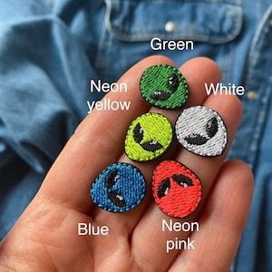 May include: Five embroidered iron-on patches in the shape of an alien head. The patches are in the colors green, neon yellow, white, blue, and neon pink.