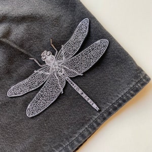 Op de afbeelding: Een zilveren geborduurde libellenpatch op een donkergrijze denim stof.