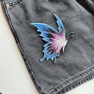 Puede incluir: Primer plano de una prenda de mezclilla gris oscuro con un parche de mariposa vibrante. El parche presenta bordados azules, morados y rosas con detalles en negro. La mezclilla tiene un lavado oscuro.