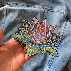 Puede incluir: Un parche bordado colorido de una flor de loto con patrones geométricos y detalles iridiscentes. La flor está bordeada con hilo dorado y tiene una forma de media luna en el centro.