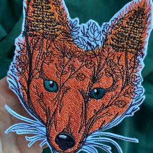 May include: Embroidered patch of a fox's face with a detailed design of foliage and flowers growing over the animal's head. The fox has a white background and orange fur with blue eyes.