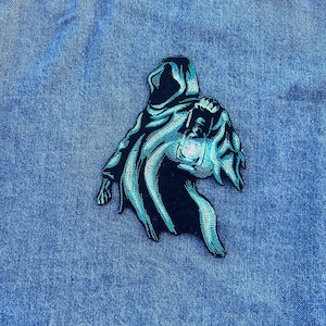May include: Embroidered patch featuring a hooded figure in black and teal. The figure is holding a lantern. The patch is on a blue denim background. The design has a detailed, textured appearance.