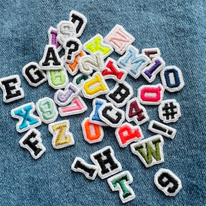 Può includere: Una collezione di toppe in tessuto ricamate con lettere, numeri e simboli in vari colori. Le toppe hanno un bordo bianco e sono sparse su uno sfondo di denim blu. Le lettere includono A, B, C, E, F, G, H, T, W, X, Y, Z.