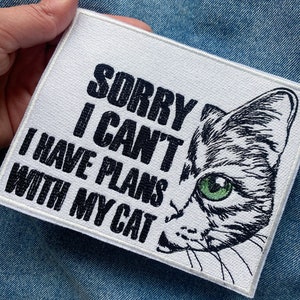 Può includere: Toppa bianca da stirare con testo nero che recita "Mi dispiace, non posso, ho progetti con il mio gatto". Un'illustrazione in bianco e nero di un viso di gatto con occhi verdi è parzialmente visibile sul lato destro della toppa.