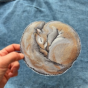 Op de afbeelding: Een ronde geborduurde patch met een eekhoorn die tot een balletje is opgerold. De eekhoorn is afgebeeld met bruine, beige en grijze draden, wat de gedetailleerde vachtstructuur en de pluizige staart rond het lichaam benadrukt.