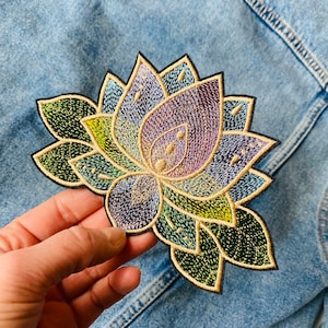 Può includere: Una toppa ricamata colorata di un fiore di loto con foglie verdi. Il fiore è un mix di viola, blu e giallo.